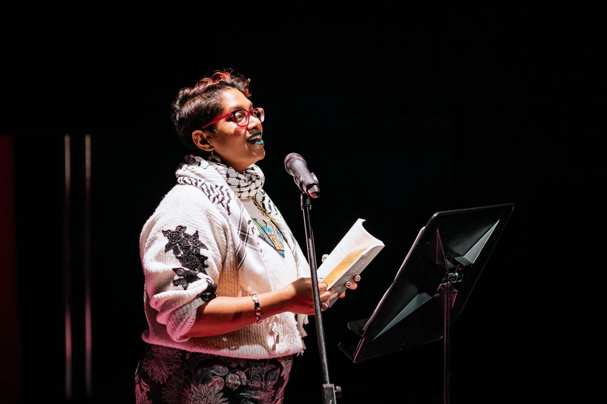 Chandrika Narayanan-Mohan, a person with short, dark hair and dark skin stands illuminated in front of a microphone, speaking while holding a book in their hands. They are wearing a light-coloured jumper and scarf featuring various patterns, and glasses. The background behind them is in darkness. In front of them is a document holder on a stand. Photo by Simon Lazewski.