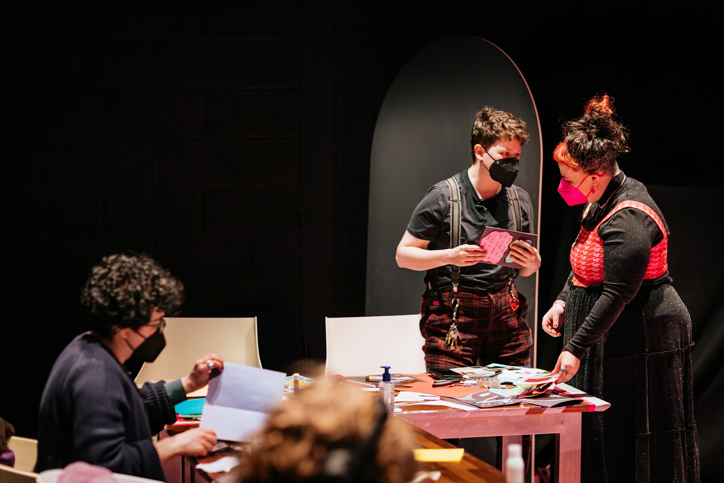 Image of artist and workshop lead Aisling Reina speaking with Disrupt festival organiser Áine O'Hara . They are pictured at a workshop, surrounded by magazine clippings. Áine wears a black dress with a pink crop top over it. Aisling wears plaid pants held up with braces, with a bundle of keys attached. Both people and a figure in the foreground wear facemasks. Photo by Simon Lazewski.