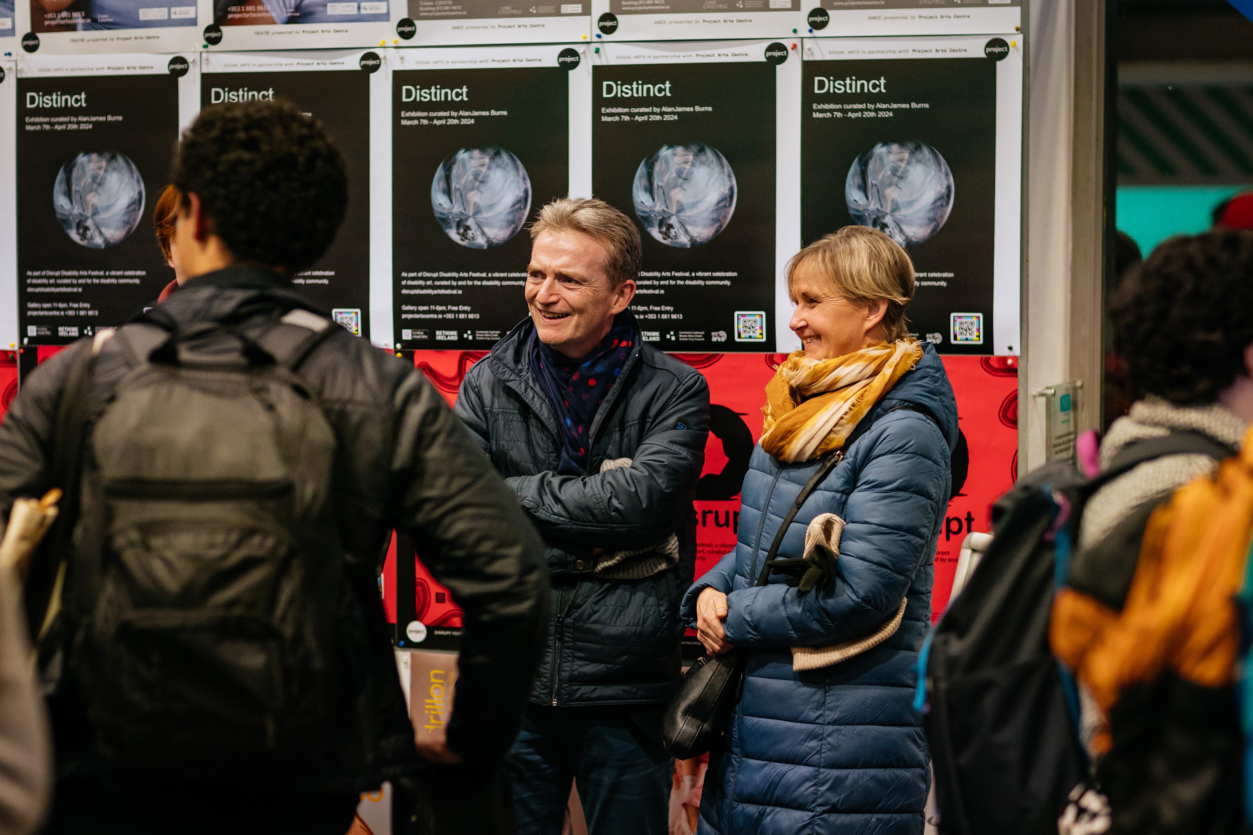 People socialise in Project Art Centre’s lobby. A masculine and a feminine figure, both dressed in dark jackets are looking at something unseen. Both have fair hair and are smiling. Behind them is a row of posters advertising Distinct, the exhibition. The posters are black and feature an icy orb, an image taken from Ruth Le Gear’s artwork. Photo by Simon Lazewski. March 9th