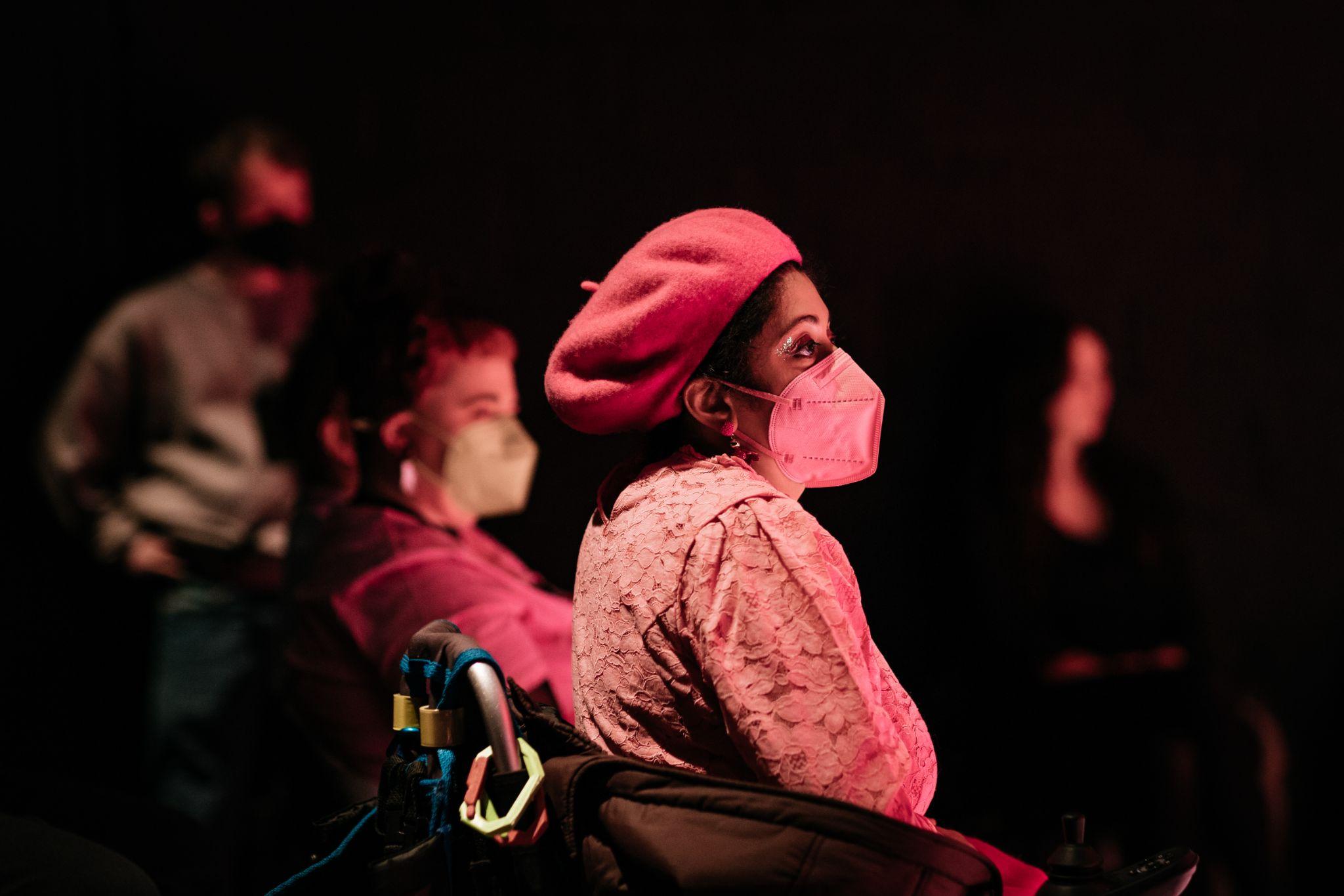 A group of people sit together, mostly at a side angle to the camera. In the foreground, a woman with dark skin and dark hair, wearing a bright pink beret, pink top, and pink mask, is visible. Beside her, slightly out of focus, is another woman wearing a pink top and mask. Two people in the background are also out of focus. The background is black. Photo by Simon Lazewski.