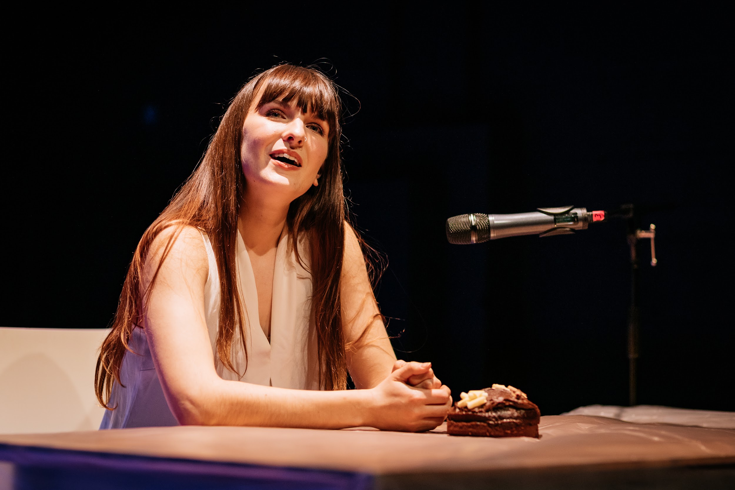 An image of artist Ali Clarke performing. She is sitting at a table with a small chocolate cake in front of her. She is well lit in bright light and looks out and up to the audience. She is a white woman with long dark hair and a fringe and wears a white sleeveless blouse. The background is completely black. There is a microphone and stands in front of Ali. Photo by Simon Lazewski.