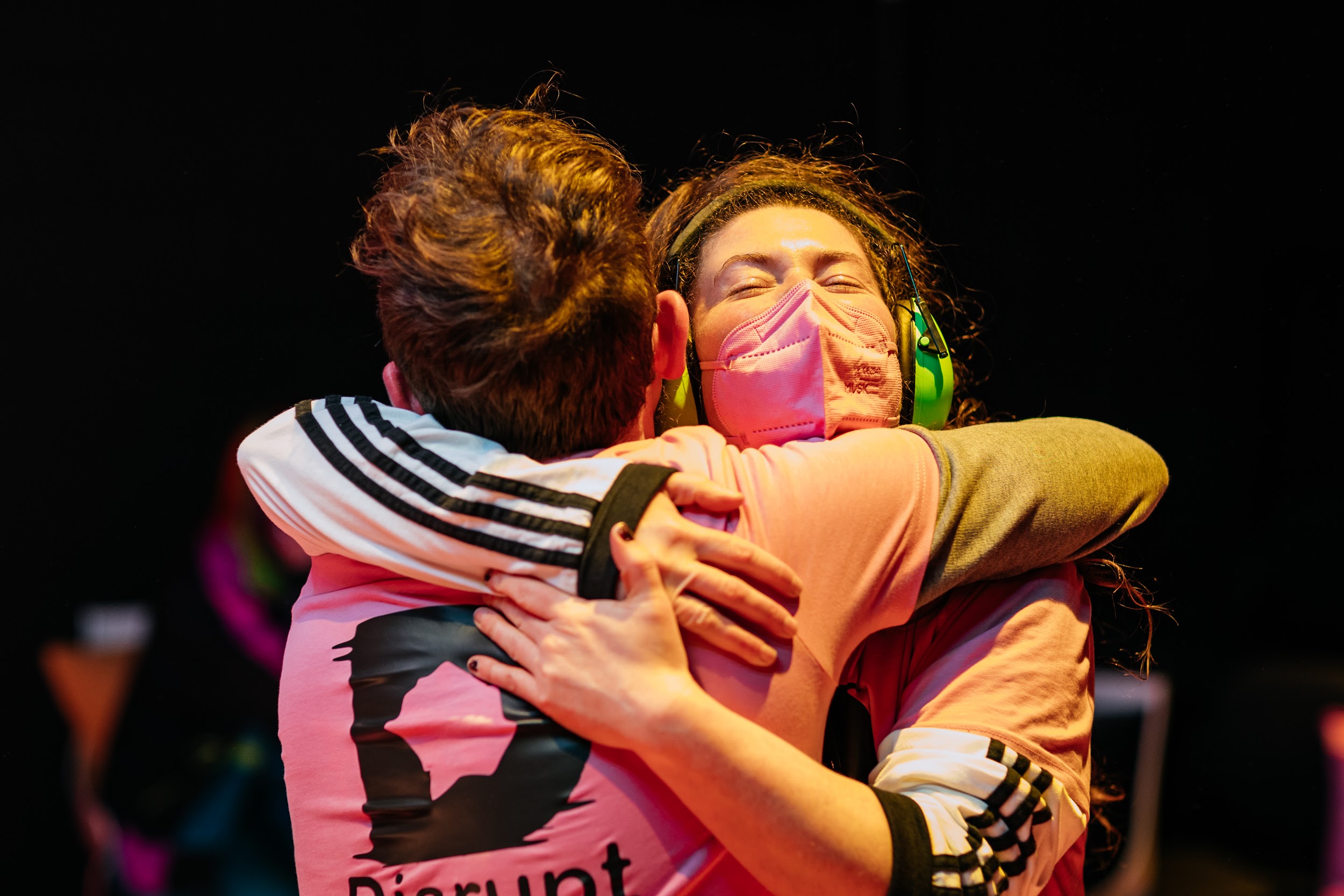 Disrupt festival organiser Alan James Burns hugs a member of crew. Alan has their back to us. The crew member, Ailbhe O’Connor, grasps Alan with both arms in an act of elation. She wears ear defenders and a pink face mask. Photo by Simon Lazewski.