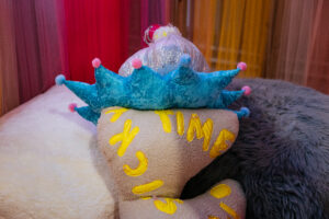 Another close shot of Rion Duffy Murphys soft sculptures. This piece has a fleece texture in an oatmeal grey, with yellow words stitched into it, the only words we can see is the word 'time'/ The cushioned piece is topped with a star like, blue crown sesction. it has several pointed parts, all topped with a pink sphere, above that the next section resembles a discoball.