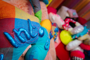 An upclose shot of the Disrupt rest area. In the foreground we see one of Rion Duffy Murphy's soft sculptures - a patchwork style large worm shaped cushion. With patches of colours in red, yellow greean and blue. We see the word 'into' in a 3d bubbled font attached to the piece. In the background we see a blurred collection of Rions pieces, colourful, comfy and inviting.