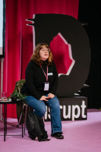 Artist and facilitator Al Bellamy leads a workshop on embedding access in production. They are sat on a chair on a pink carpetted stage, engaging in discussion with the audience.
