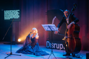Artist Emilie Conway sits on the floor while singing. The scence is of warm purples and reds and a spotlight behind Emilie illuminates her warmly. Shes wearing a flowy long ballgown and to her right we see a suited man playing the cello.