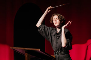 Artist Day Magee deep in their performance. They wear all black and stand at a lecturn. Eyes closed, they hold a wand like instrument in their hand above there head, the other softly gesturing to their face. 