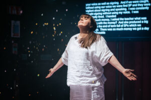Artist Lianne Quigley during the peformance of her show 'A Softer Kiss'. Lianne wears an all white linen outfit, has their arms outstretched towards the ceiling and has her eyes clothes. There is gold confetti falling from the sky and its a magical scene.