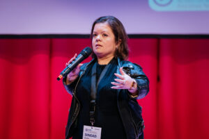 Sinead Burke, Activist and CEO of Tilting the Lens, addresses an audience from the stage, hand gesturing in conversation. She wears all black, mic in hand and behind her we see a curtained wall and a presentation screen. 