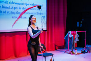 Artist Porcelain Delaney smiles broadly towards the audience. In one hand she holds a cane, the other she swings overhead a pompom like fabric. Behind Porcelain we see captions on a screen and in the background a railing of costume changes.