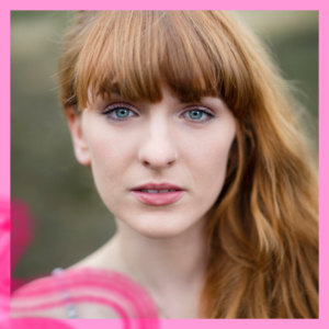 Alison Clarke looks straight at the camera, red hair swept over one shoulder, blue eyes below her fringe. Behind her we see a blurred landscape of green.
