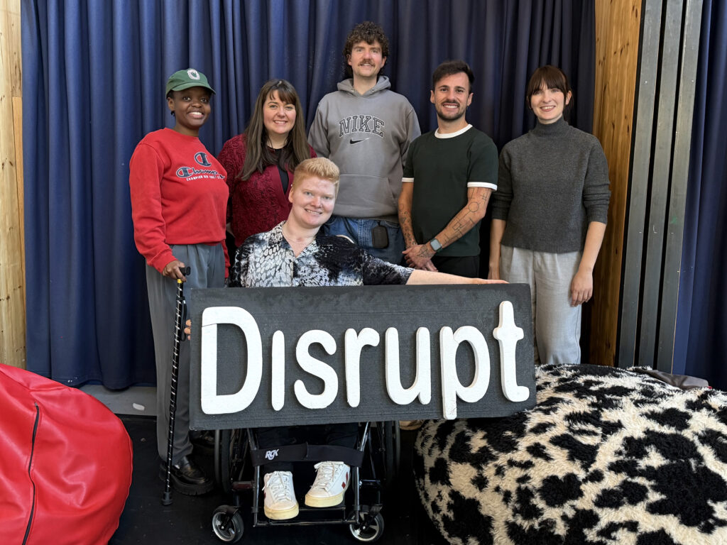 6 people pose for a group photo. They are all smiling warmly, standing against a navy curtained wall. In front of them we see 2 large bean bags, one red and one black and white animal print. Standing in the back row, on the left we have Tino Wekare with short hair under a green hat and in their right hand hold onto a cane. Next we have Lianne Quigley, long brown hair and wearing a berry colour cardigan. Next Tadhg Kinsella standing the talest in the line, with a grey hoodie, short curly hair and a moustache. Next Omar Lorenzo García wearing a green tshit and tattoed arms crossed in front, they have short dark hair and a short beard. Next Alison Clarke, wearing a grey knitted jumper, brown hair tied up with a fringe. In the front Ciara O'Mahony, short blonde hair, holding onto the large 'Disrupt' 3D sign. Ciara holds one arm out holding onto the sign and rests it upon her wheelchair.